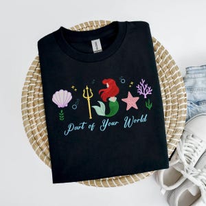 May include: Black t-shirt with an embroidered design of a mermaid, seashell, starfish, and coral. The text "Part of Your World" is written in a script font. The shirt is displayed on a woven basket.