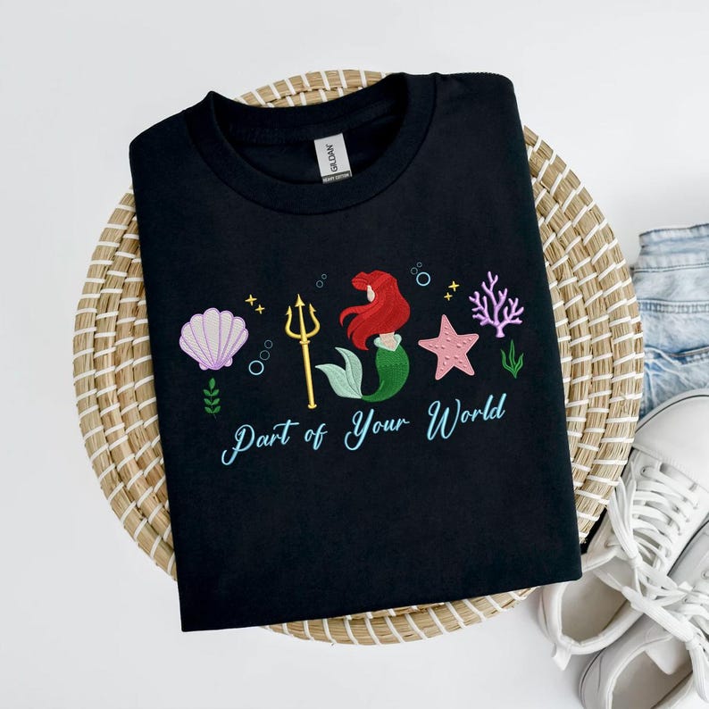 May include: Black t-shirt with an embroidered design of a mermaid, seashell, starfish, and coral. The text "Part of Your World" is written in a script font. The shirt is displayed on a woven basket.