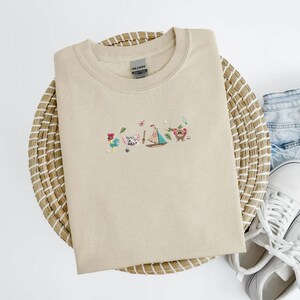May include: Beige crewneck sweatshirt with embroidered cartoon characters, including a rooster, pig, sailboat, and a fruit-like figure. The sweatshirt is folded on a woven basket, with denim shorts and white sneakers visible in the background.