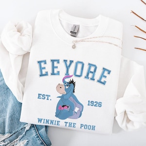 May include: White sweatshirt featuring an embroidered Eeyore design from Winnie the Pooh. The design includes the word "Eeyore" in blue block letters, the character Eeyore, and the text "EST. 1926" and "WINNIE THE POOH". A silver necklace is draped across the sweatshirt.