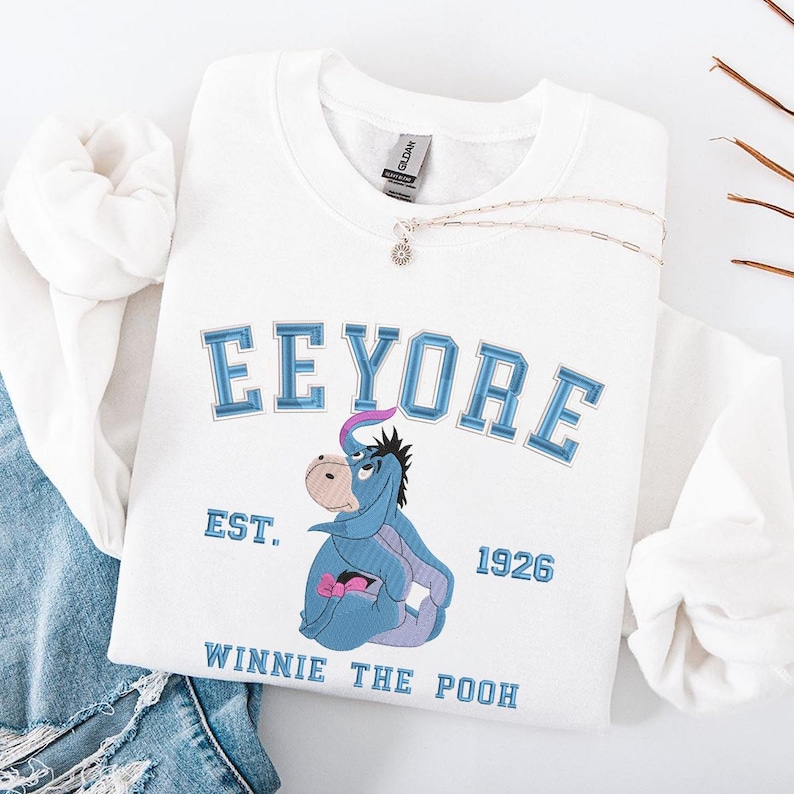 May include: White sweatshirt featuring an embroidered Eeyore design from Winnie the Pooh. The design includes the word "Eeyore" in blue block letters, the character Eeyore, and the text "EST. 1926" and "WINNIE THE POOH". A silver necklace is draped across the sweatshirt.