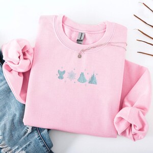 May include: A light pink sweatshirt with embroidered light blue and white designs of a bird, snowflake, dress, and castle. A silver necklace with a sun pendant rests on the sweatshirt. The sweatshirt is folded with a pair of blue jeans.