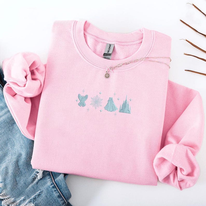May include: A light pink sweatshirt with embroidered light blue and white designs of a bird, snowflake, dress, and castle. A silver necklace with a sun pendant rests on the sweatshirt. The sweatshirt is folded with a pair of blue jeans.