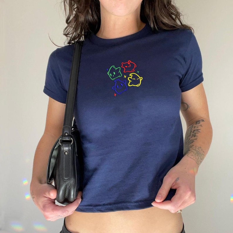 May include: Navy blue t-shirt with a crew neck and short sleeves. The shirt features a colorful embroidered design of four cartoon faces. A black leather shoulder bag is also visible.