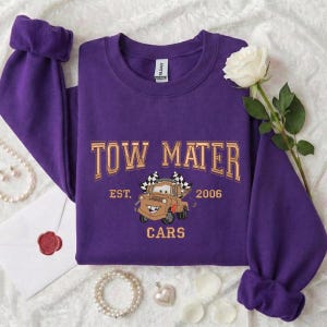 Puede incluir: Una sudadera morada con la inscripci&oacute;n "TOW MATER" en letras doradas, junto con una imagen de Mate de la pel&iacute;cula Cars. La sudadera tambi&eacute;n presenta "EST. 2006" y "CARS".
