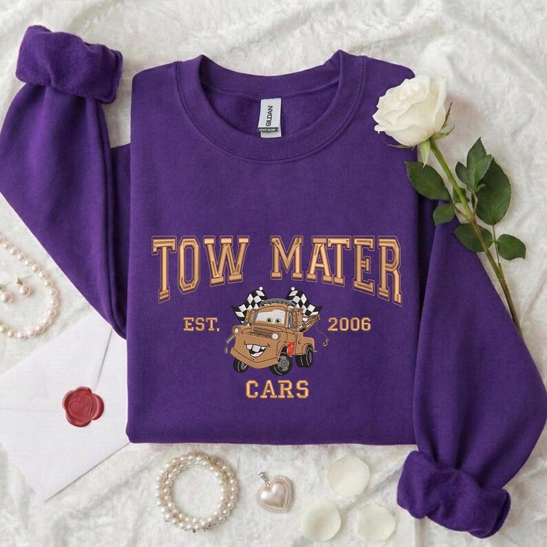 Puede incluir: Una sudadera morada con la inscripci&oacute;n "TOW MATER" en letras doradas, junto con una imagen de Mate de la pel&iacute;cula Cars. La sudadera tambi&eacute;n presenta "EST. 2006" y "CARS".