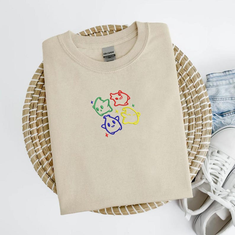 May include: A beige crew neck sweatshirt with an embroidered design of four colorful cartoon ghosts. The ghosts are outlined in green, red, blue, and yellow, with small star accents. The sweatshirt is folded on a woven basket.