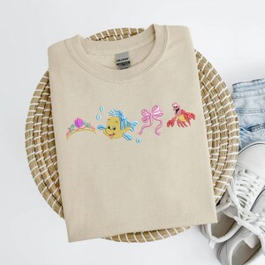 May include: Beige crewneck sweatshirt featuring embroidered cartoon characters. The design includes a tiara, a yellow fish with blue fins, a pink bow, and a red crab. The sweatshirt is folded on a woven mat with denim shorts and white sneakers.