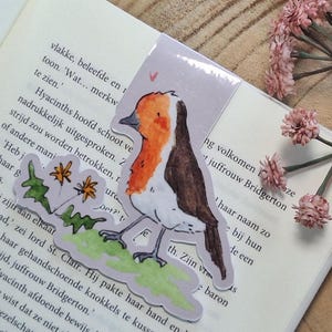 Peut inclure: Un marque-page avec une illustration à l'aquarelle d'un rouge-gorge perché sur une branche, sur un livre ouvert et des clés antiques. Le marque-page est posé sur une surface en bois avec des accents floraux roses.