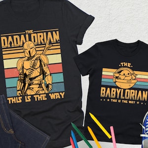 以下が含まれることがあります： スターウォーズをテーマにしたグラフィックがプリントされた黒いTシャツ2枚。1枚はマンダロリアンのイラストと「The Dadalorian」、もう1枚はグローグーのイラストと「The Babylorian」の文字。どちらのシャツにも「This is the Way」と書かれています。青いジーンズと青いスニーカーも写っています。