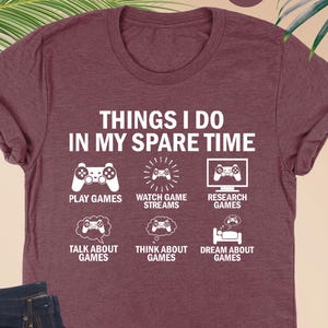 May include: A heather maroon short-sleeve t-shirt featuring white text that reads THINGS I DO IN MY SPARE TIME, accompanied by six icons representing gaming activities including playing, watching, researching, talking, thinking, and dreaming.