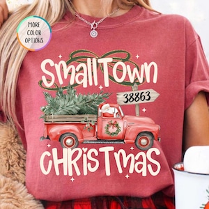 May include: A crimson t-shirt featuring a graphic of a red pickup truck carrying a green Christmas tree. The text reads Small Town CHRISTMAS and 38863 on a sign. A small circular badge says MORE COLOR OPTIONS and a tag says CRIMSON.