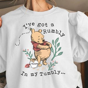 May include: A white crewneck sweatshirt featuring a graphic of Winnie the Pooh holding a honey pot with the text I've got a Rumbly In my Tumbly, accompanied by a small bee, a ladybug, and green leafy sprigs printed on the front center.