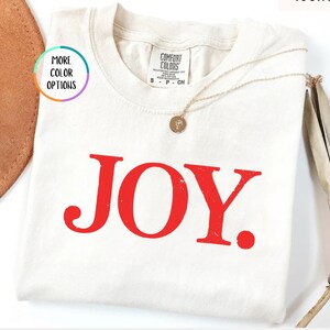 May include: An ivory short-sleeved t-shirt featuring the word JOY. printed in bold red capital letters across the chest. The shirt has a Comfort Colors brand label at the inner neckline and a thin gold-toned necklace resting on the fabric.