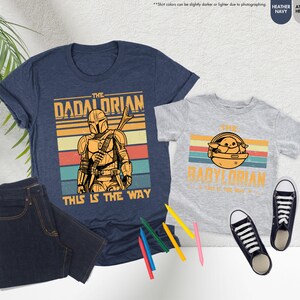 以下が含まれることがあります： 「The Dadalorian」と「The Babylorian」のグラフィックがプリントされた、ネイビーブルーとヘザーグレーのTシャツ。濃い色のジーンズ、白と黒のスニーカー、色鉛筆も画像に写っています。