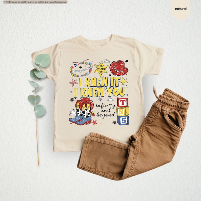 Puede incluir: Una camiseta de color natural con un estampado gr&aacute;fico que incluye el texto I KNEW IT I KNEW YOU infinity and beyond. El dise&ntilde;o presenta una insignia de sheriff, un sombrero de vaquero rojo, botas vaqueras y bloques de letras T, S y 5.