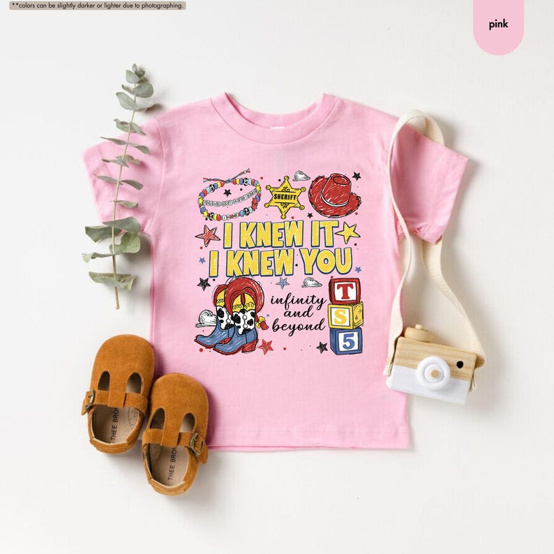 Puede incluir: Una camiseta rosa claro de manga corta con un estampado gr&aacute;fico que incluye el texto I KNEW IT I KNEW YOU infinity and beyond. El dise&ntilde;o muestra una estrella de sheriff, un sombrero de vaquero rojo, botas y bloques con las letras T, S y 5.