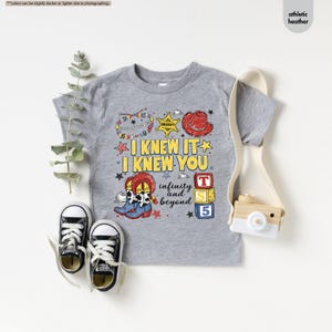 Puede incluir: Una camiseta gris jaspeado atl&eacute;tico con un estampado gr&aacute;fico que incluye el texto I KNEW IT I KNEW YOU infinity and beyond. El dise&ntilde;o presenta una estrella de sheriff, un sombrero de vaquero rojo, botas con estampado de vaca y bloques de colores con el n&uacute;mero 5.
