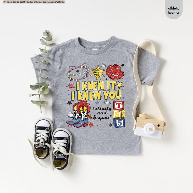 Puede incluir: Una camiseta gris jaspeado atl&eacute;tico con un estampado gr&aacute;fico que incluye el texto I KNEW IT I KNEW YOU infinity and beyond. El dise&ntilde;o presenta una estrella de sheriff, un sombrero de vaquero rojo, botas con estampado de vaca y bloques de colores con el n&uacute;mero 5.