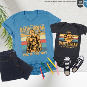 以下が含まれることがあります： 「The Dadalorian」のグラフィックが入った水色のTシャツと、「The Babylorian」のグラフィックが入った濃いグレーのベビーボディスーツ。どちらも、キャラクターのイラストと「This is the Way」というフレーズが入ったレトロなデザインです。濃い青色のジーンズと黒いスニーカーも画像にあります。
