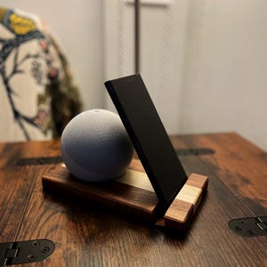 May include: A wooden stand with a black phone holder and a gray smart speaker. The stand is made of dark and light wood. The phone holder is angled, and the speaker is round. The stand is on a dark wood table.