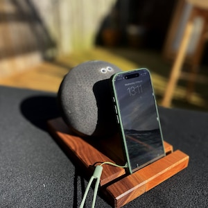 May include: A wooden phone stand with a dark finish holds a smartphone with a green case. A gray, spherical speaker rests on the stand. A green lanyard is attached to the stand. The phone displays the time: 13:11.