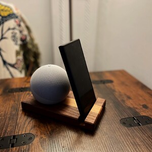 May include: A wooden phone stand with a black phone and a gray spherical speaker on a dark wood table. The stand is made of two types of wood, with a lighter wood accent. The table has metal hinges.