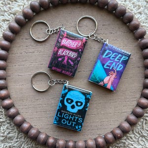 May include: Three miniature book keychains with silver key rings. The book covers feature the titles "Butcher & Blackbird", "Deep End", and "Lights Out". The keychains are arranged on a wooden tray with a beaded border, set against a neutral background.