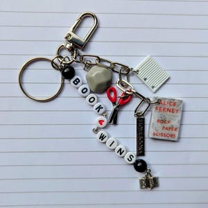 May include: A silver keychain with various charms, including a gray rock, red scissors, and a miniature book. The keychain spells out "BOOK WINS" with letter beads and includes a small book charm with the text "ALICE FEENEY ROCK PAPER SCISSORS".
