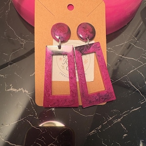 May include: A pair of handmade earrings with a rectangular shape and a circular top. The earrings are a vibrant magenta color with a marbled effect. They are attached to a small, tan card with a logo.