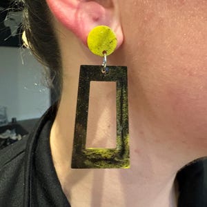 May include: A pair of earrings featuring a yellow circular stud and a black rectangular drop with a yellow interior. The earrings are connected by a silver-colored metal loop. The earrings are a bold statement piece.