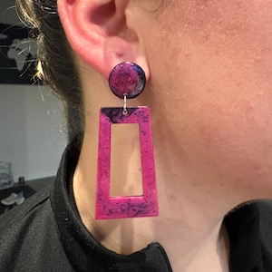 May include: Earrings featuring a circular stud and a rectangular drop. The earrings are a bright pink with dark purple and black marbling. They are attached with a silver-coloured metal hook.