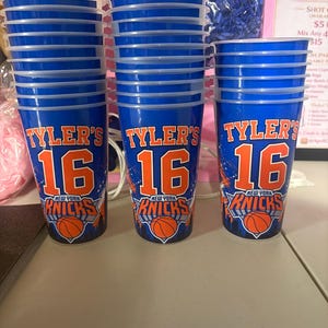May include: Three blue plastic cups stacked in columns, each featuring the text 'TYLER'S 16' in orange and white lettering above a New York Knicks basketball team logo with an orange basketball graphic at the base of each cup.