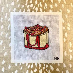 May include: Needlepoint canvas featuring a beige tote bag filled with hot dogs. The hot dogs have red and yellow toppings. The tote bag has red handles and trim. The background is a white canvas with a dotted pattern.