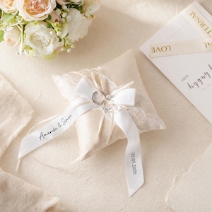 May include: A square cream-coloured ring pillow featuring lace trim and a white satin ribbon tied in a bow. The ribbon is printed with the names Amanda & Simon and the date 02.05.2026. Two silver rings are secured to the centre of the bow.