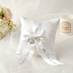 May include: A square white satin ring pillow featuring a central white ribbon tied in a bow. Two silver-toned rings are secured by the ribbon. The ribbon is printed with the black text Paul & Chloe and 15th February 2027 in a script font.