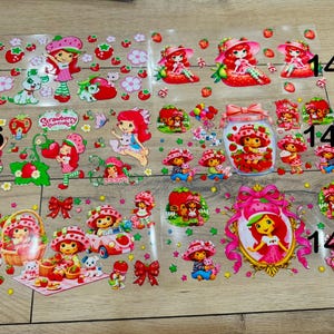 May include: A collection of six transparent sticker sheets featuring Strawberry Shortcake characters, strawberries, and bows, labeled with numbers 144 through 149. The designs include various scenes of the character with pets, in a car, and in a frame.