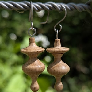 Puede incluir: Un par de pendientes de madera hechos a mano con un diseño simétrico único. Cada pendiente presenta una forma de madera torneada de color marrón claro, con un centro bulboso, una parte superior ensanchada y una base en forma de lágrima. Cuelgan de ganchos plateados, sobre un fondo verde borroso.