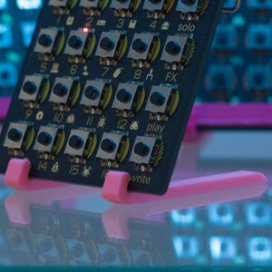 Könnte beinhalten: Eine schwarze elektronische Leiterplatte mit einem Raster aus quadratischen Drucktasten und gelben kreisförmigen Akzenten. Die Platine zeigt nummerierte Beschriftungen von 1 bis 16 sowie Symbole und Text, darunter solo, FX, play und write. Sie steht auf einem rosa Ständer.