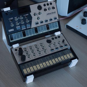 Könnte beinhalten: Zwei Korg Volca Synthesizer, das Volca Drum und das Volca Keys, sind auf einem weißen, zweistufigen Ständer montiert. Die Geräte verfügen über schwarze und silberne Bedienfelder mit verschiedenen Reglern, Tasten und einer Touch-Tastatur.