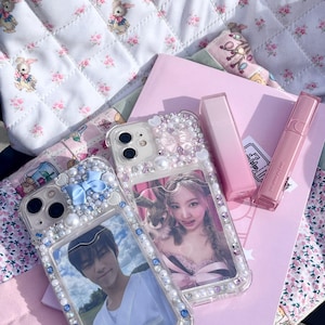 May include: Two clear phone cases decorated with pearls and rhinestones, each holding a photo. One case has a blue bow. Pink lipsticks and a pink notebook are also visible, along with a floral patterned bag.