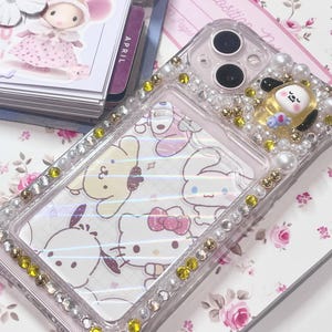 Puede incluir: Funda de teléfono transparente decorada con pedrería, perlas y detalles dorados. La funda presenta un diseño con personajes de dibujos animados. Un pequeño personaje amarillo está en la parte superior derecha. La funda está sobre un fondo floral.