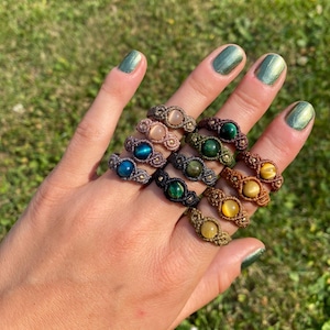 Könnte beinhalten: Eine Sammlung von Makramee-Ringen mit runden Cabochon-Edelsteinen in verschiedenen Farben wie Blau, Grün, Gelb und Pfirsich. Jeder Ring ist mit aufwendiger Knotentechnik gefertigt und präsentiert einen einzigartigen Stein in der Mitte des Bandes.