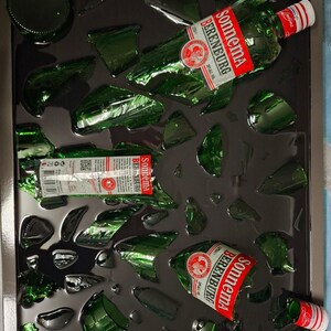 Op de afbeelding: Een rechthoekig ingelijst kunstwerk met verspreide scherven groen glas en fragmenten van Sonnema Berenburg flessen. Het display bevat flessenhalzen met rood-witte etiketten, een bodem en diverse glasstukjes tegen een donkere achtergrond.