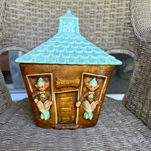 May include: A vintage ceramic cookie jar shaped like a whimsical house. The house features a brown body, a turquoise roof, and two elf figures. The jar has a door and windows, with the word "Sweet" above the door.