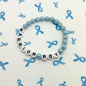 May include: A beaded bracelet with the word "WARRIOR" spelled out in white letter beads. The bracelet features light blue pearl beads and silver accents. The background is white with blue ribbon graphics.