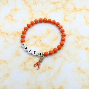 May include: Orange beaded bracelet with the word "FAITH" spelled out in white letter beads. A silver charm with an orange ribbon hangs from the bracelet. The bracelet is on a marble-patterned surface.