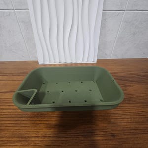 Peut inclure: Un bac à plantes rectangulaire vert olive avec des trous de drainage et un compartiment latéral triangulaire. Un panneau mural blanc à motifs ondulés est en arrière-plan. Le bac repose sur une surface en bois.