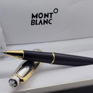 Può includere: Una penna a sfera Montblanc nera con finiture dorate, presentata in una scatola bianca. La penna è parzialmente aperta, rivelando un pennino argentato e dorato. Un sacchetto di gel di silice è visibile.