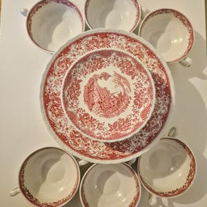 May include: A set of white ceramic dinnerware featuring a red transferware pattern. The collection includes six teacups with red floral rims and two stacked plates, with the smaller top plate displaying a detailed red landscape scene.